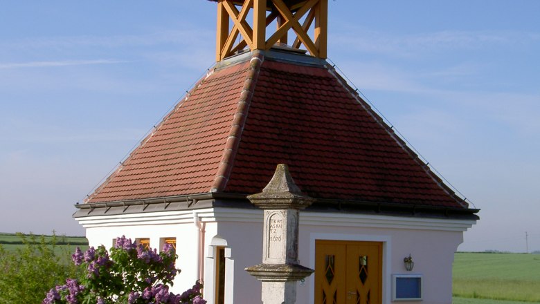 Kleine Kapelle mit rotem Dach und Kreuz, umgeben von Blumen und einer Bank, bei klarem Himmel.