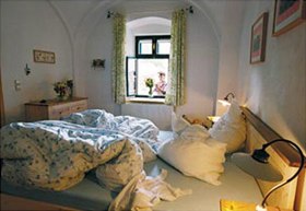 Bilderbuch-Bauernhof, © Michael Himml Gemütliches Schlafzimmer mit ungemachtem Bett und Blick aus dem Fenster auf eine Person mit Blumen.