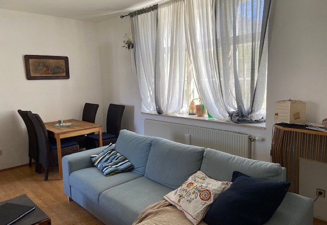 Ein Wohnzimmer mit einem blauen Sofa, einem Holztisch mit St&uuml;hlen und einem Fenster mit wei&szlig;en Vorh&auml;ngen.