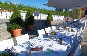 Langer, festlich gedeckter Tisch im Freien mit weißen Tischdecken und Stühlen, umgeben von Pflanzen und unter einem Sonnensegel.