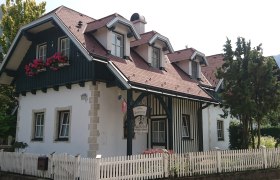 Ein traditionelles Landhaus mit Holzverkleidung, roten Dachziegeln und Blumenkästen vor den Fenstern. Ein weißer Holzzaun umgibt das Grundstück.