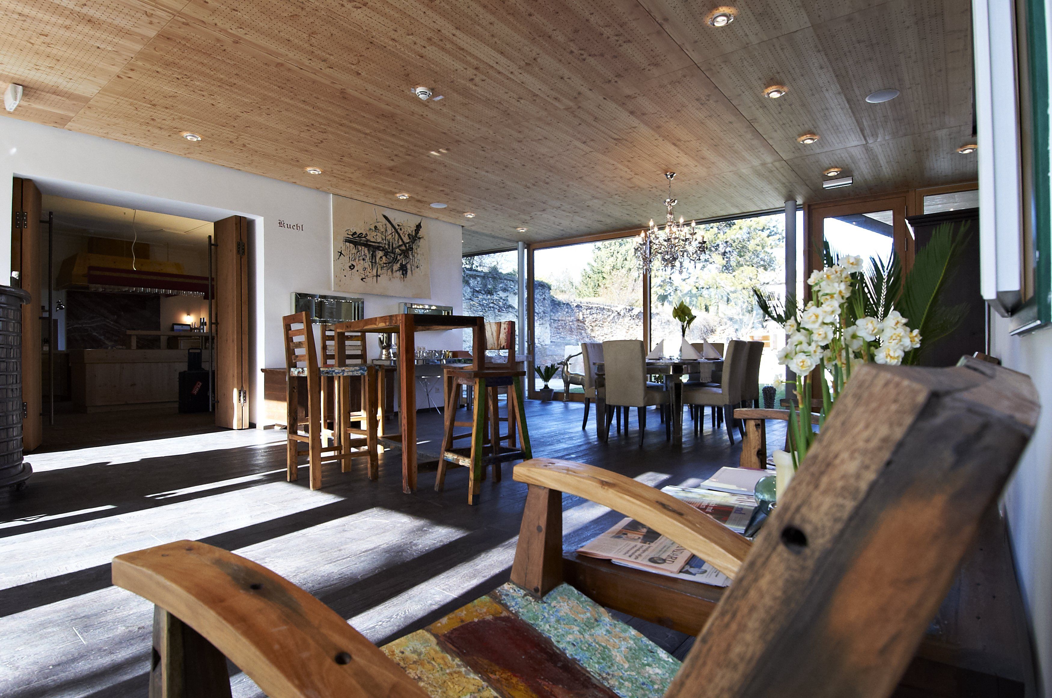 Ein moderner Frühstücksraum mit Holzmöbeln, großen Fenstern und einem Kronleuchter. Im Vordergrund ein Holzstuhl, im Hintergrund ein Esstisch.