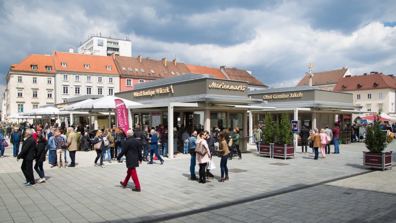 Menschenmenge auf einem belebten Marktplatz mit Ständen und Gebäuden im Hintergrund.