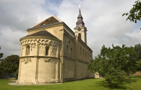Romanische Kirche mit Verzierungen und Turm, umgeben von gr&uuml;ner Wiese und B&auml;umen.