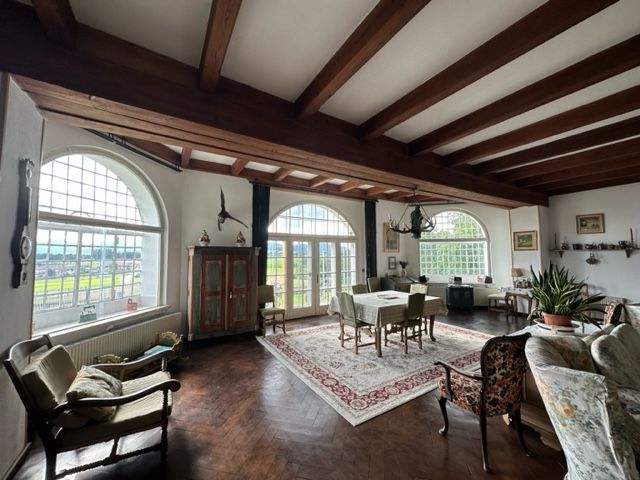 Ein elegantes Wohnzimmer mit Holzbalkendecke, großen Fenstern, antiken Möbeln und einem Teppich.