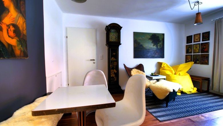 Wohnzimmer mit Tisch, St&uuml;hlen, Standuhr, Gem&auml;lden und gelbem Sitzsack.