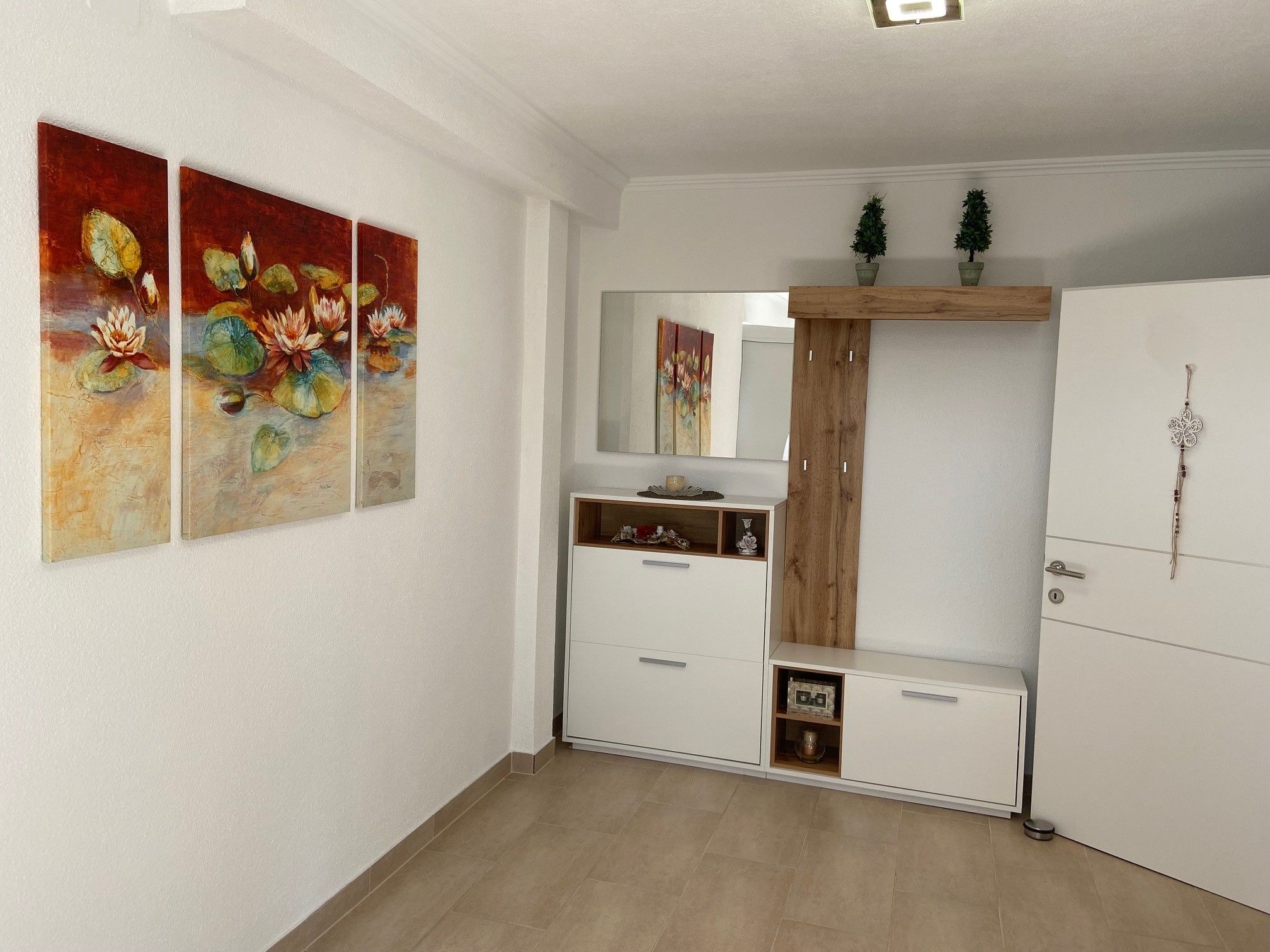 Ein Vorraum mit einem dreiteiligen Seerosenbild an der Wand, einem weißen Schrank mit Spiegel und Dekorationen.