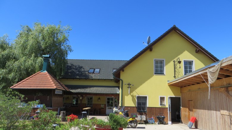 Gasthof Oase am Teich, © Urani Gelbes Gebäude mit grünem Dach und Terrasse, umgeben von Pflanzen, unter blauem Himmel.