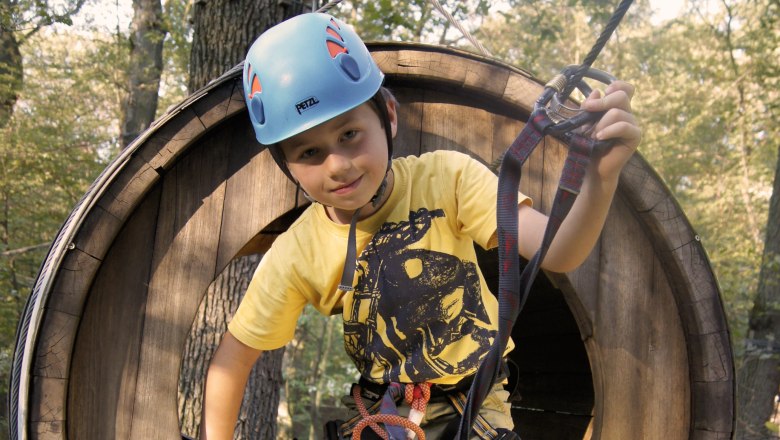 Ein Kind mit Helm und Kletterausr&uuml;stung in einem h&ouml;lzernen Kletterpark-Tunnel.