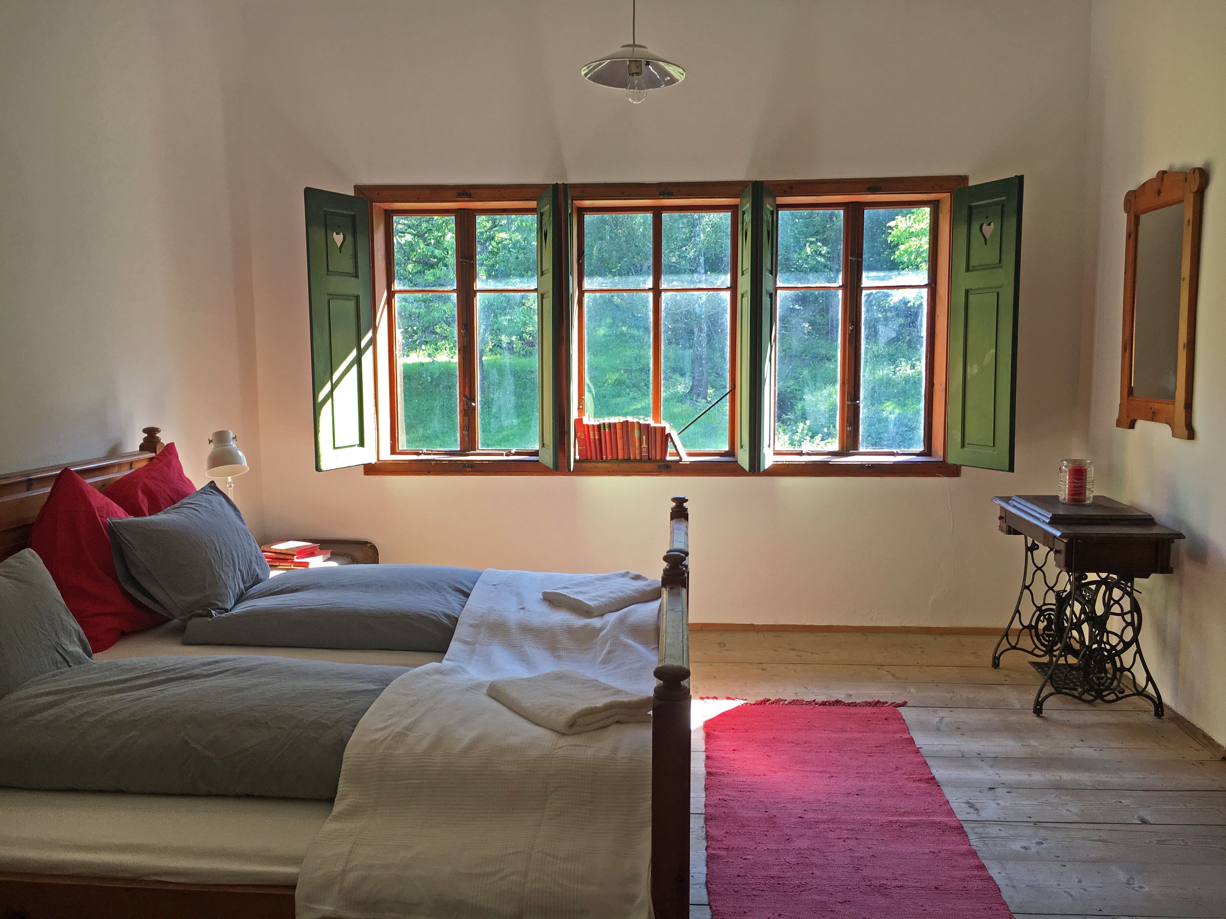 Gemütliches Doppelzimmer mit Holzboden, Bett, Fenster mit grünen Fensterläden und einem roten Teppich.