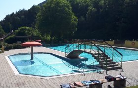 Ein Freibad mit einem kleinen Pool, einer Brücke und einem Pilz-Wasserspiel. Umgeben von Bäumen und einem sonnigen Himmel.