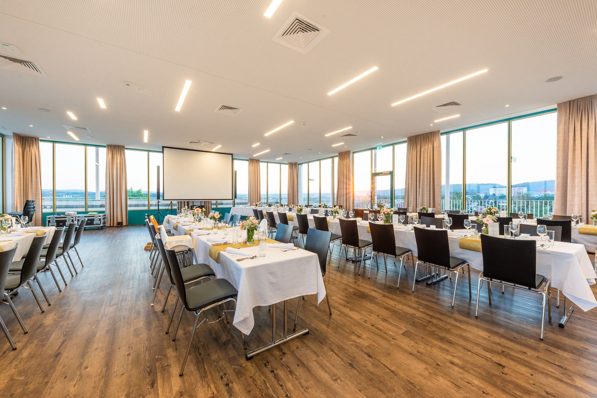 Ein großer, heller Saal mit gedeckten Tischen und Stühlen, großen Fenstern und einem Projektor.