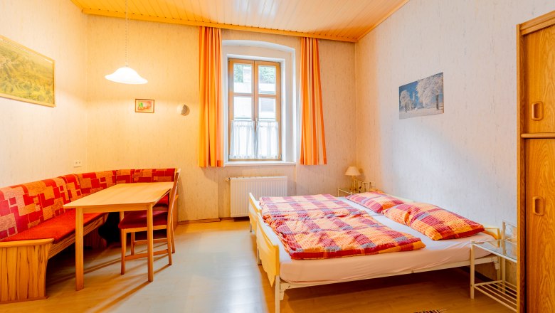 Gemütliches Schlafzimmer mit Doppelbett, Holzmöbeln und Esstisch. Helle Vorhänge und warme Farben dominieren den Raum.