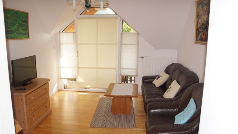 Wohnzimmer mit Ledersofa, Holztisch, Kommode und Fernseher. Fenster mit Jalousien, Bilder an den W&auml;nden.