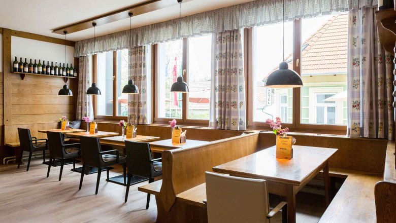 Gemütliches Gastzimmer mit Holztischen, Stühlen und Bänken, dekoriert mit Blumen und Speisekarten. Große Fenster mit Vorhängen und hängenden Lampen.