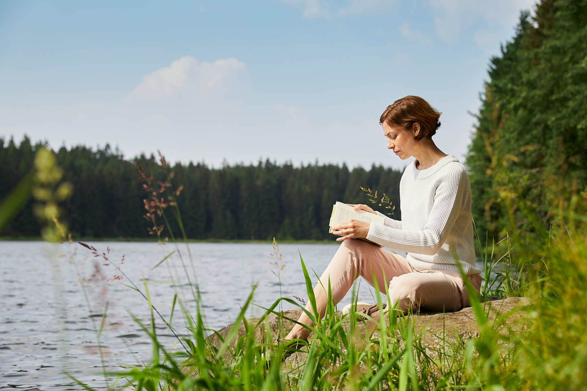Am Ufer eines ruhigen Sees sitzt eine Frau entspannt und vertieft in ein Buch. Umgeben von üppigem Grün und dem sanften Plätschern des Wassers, strahlt die Szenerie eine friedliche Atmosphäre aus, die zum Verweilen einlädt.