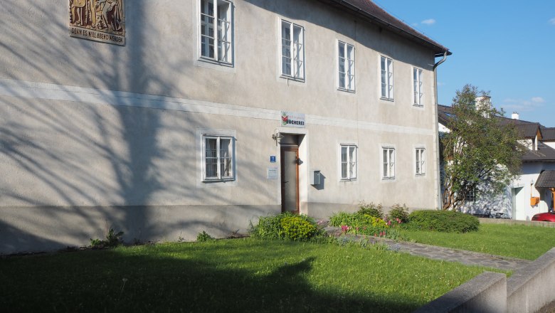 Ein zweistöckiges Gebäude mit mehreren Fenstern und einem Schild über der Tür, umgeben von einem gepflegten Garten.