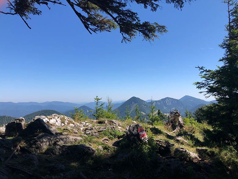 Berglandschaft mit Felsen und Bäumen im Vordergrund, blauer Himmel im Hintergrund.