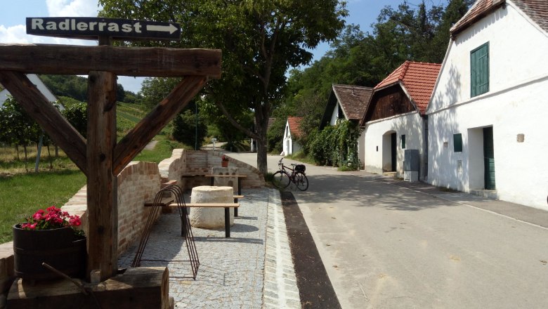 Ein Rastplatz f&uuml;r Radfahrer in Mailberg mit einem Holzschild, das auf die Radlerrast hinweist. Im Hintergrund sind Weinkeller und ein Fahrrad zu sehen.