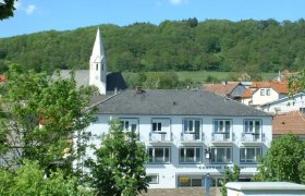 Ein zweistöckiges Gebäude mit der Aufschrift 'Gasthof Schauer' vor einem bewaldeten Hügel und einer Kirche mit spitzem Turm.