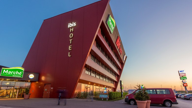 Ibis Vienna Airport, © Marché Restaurants Österreich GmbH Modernes Hotelgebäude mit schräger Fassade und Leuchtschrift 'ibis HOTEL'.