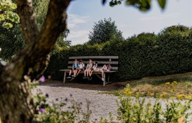 Vier Kinder sitzen auf einer &uuml;bergro&szlig;en Bank in einem Garten, umgeben von B&auml;umen und Str&auml;uchern.