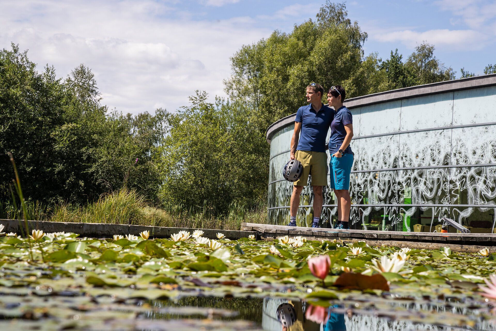 Inmitten einer malerischen Wasserlandschaft stehen zwei Radfahrer, die die Ruhe und Schönheit der Natur genießen. Umgeben von üppigem Grün und blühenden Seerosen spiegelt sich der Himmel im klaren Wasser, während die sanfte Brise eine erfrischende Atmosphäre schafft.