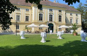 Außenansicht Schloss, im Vordergrund eine Wiese mit weißen Stehtischen und weißen Sonnenschirmen