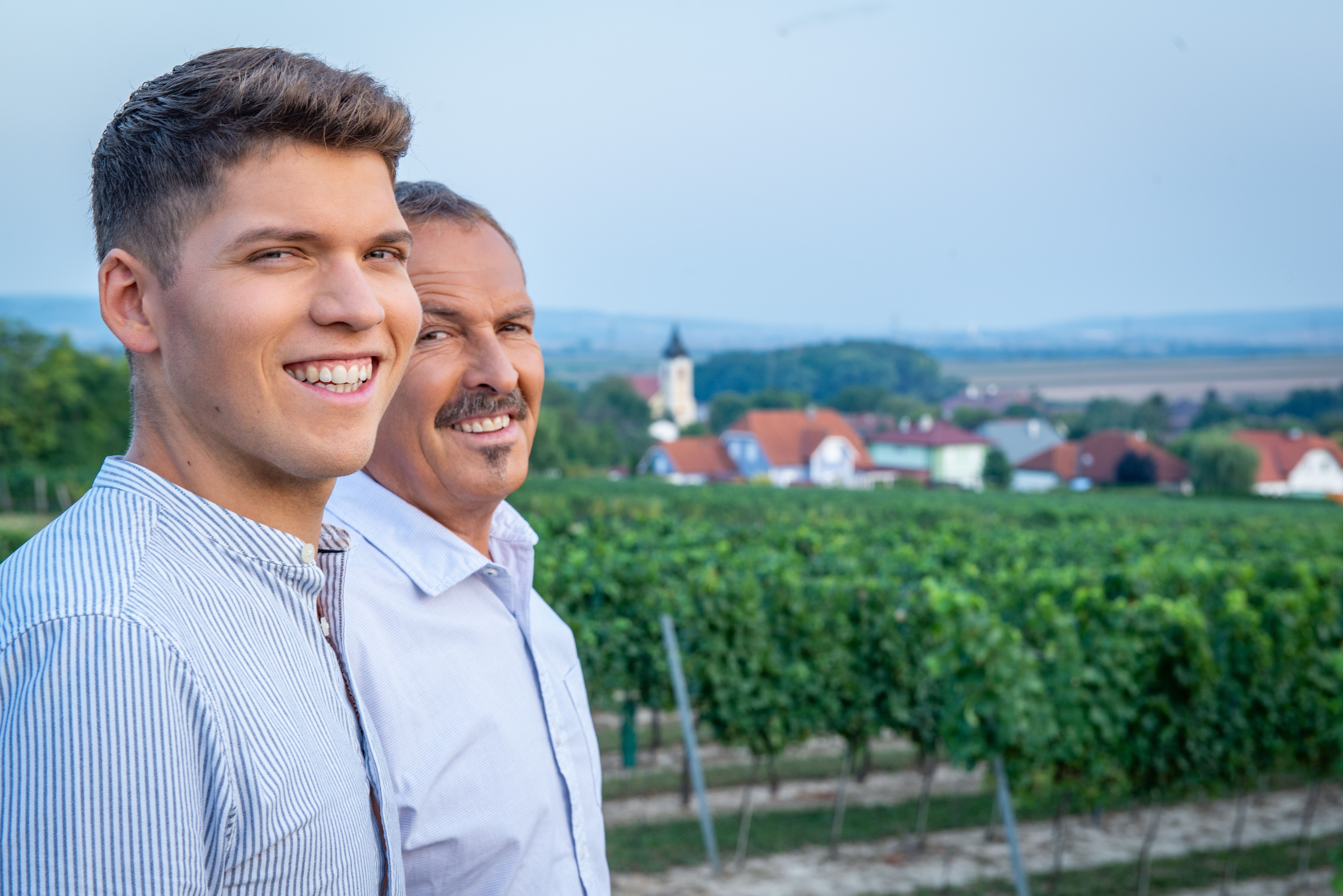 Zwei Männer lächeln in einem Weinberg mit Dorf im Hintergrund.