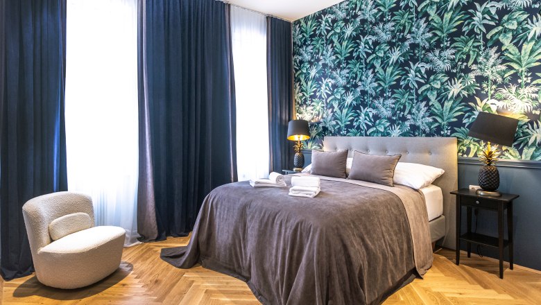 Modernes Doppelzimmer mit grauem Bett, tropischer Tapete und dunklen Vorh&auml;ngen.