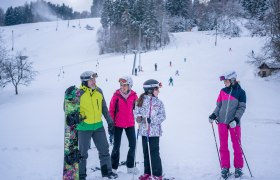 Gruppe von Skifahrern und Snowboardern auf einer verschneiten Piste mit Wald im Hintergrund.