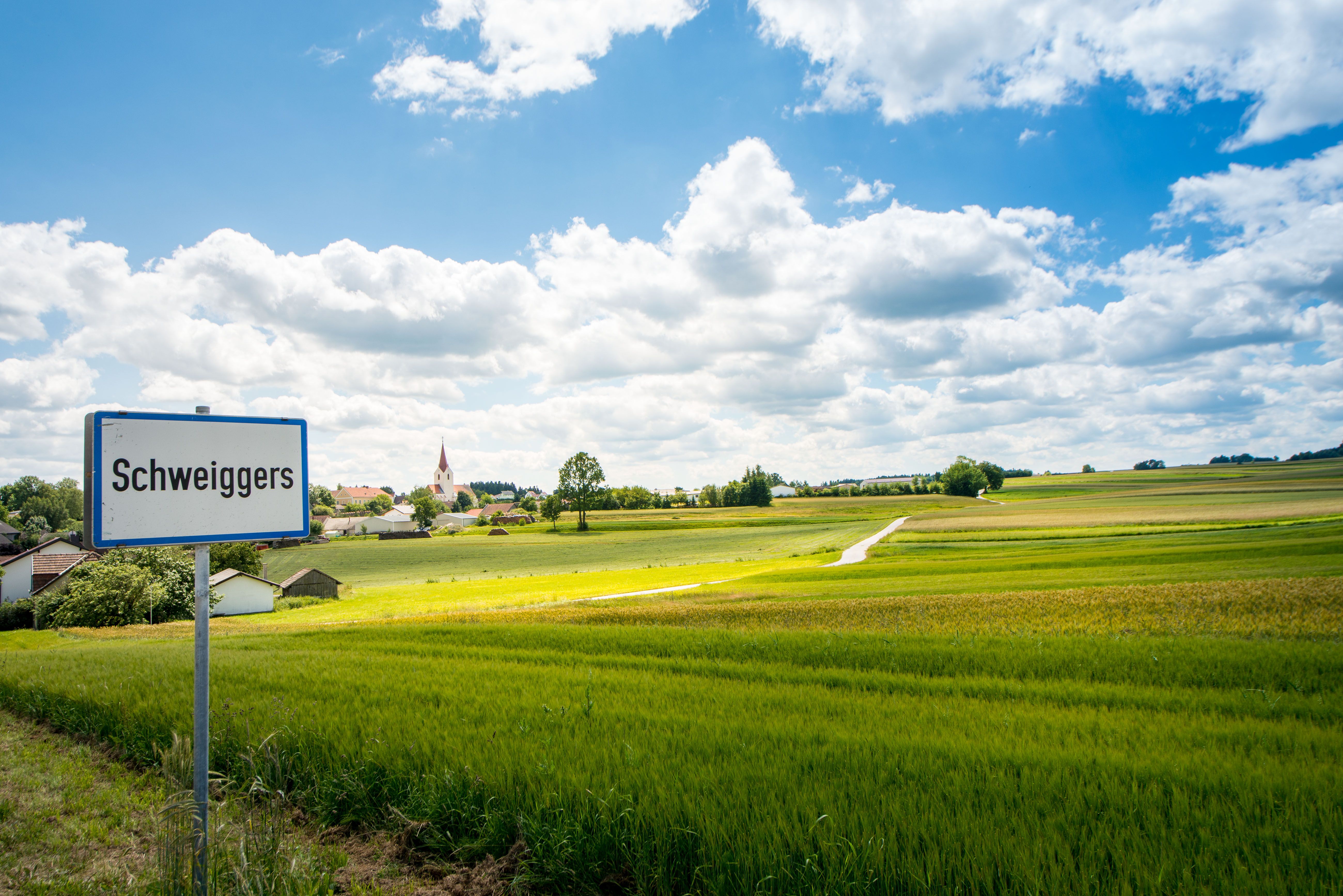 Ortstafel Schweiggers mit Blick auf grüne Felder und Häuser