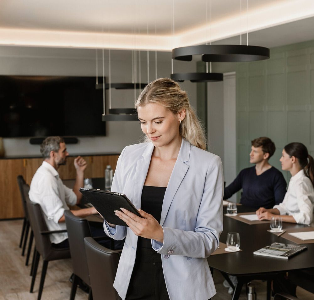Eine Frau im Vordergrund am Tablet, drei Personen in einem Business Meeting in modernem Setting an einem schwarzen Konferenztisch im Mörwald Gutshof.