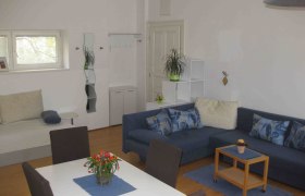 Apartment mit blauen Sofa, weisem Tisch und blauen Stühlen. Im Hintergrund ein kleines Fenster, Tür und Spiegel.