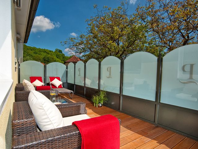 Eine Terrasse mit Rattanmöbeln, roten Decken und Kissen, umgeben von Glaswänden und Bäumen im Hintergrund.