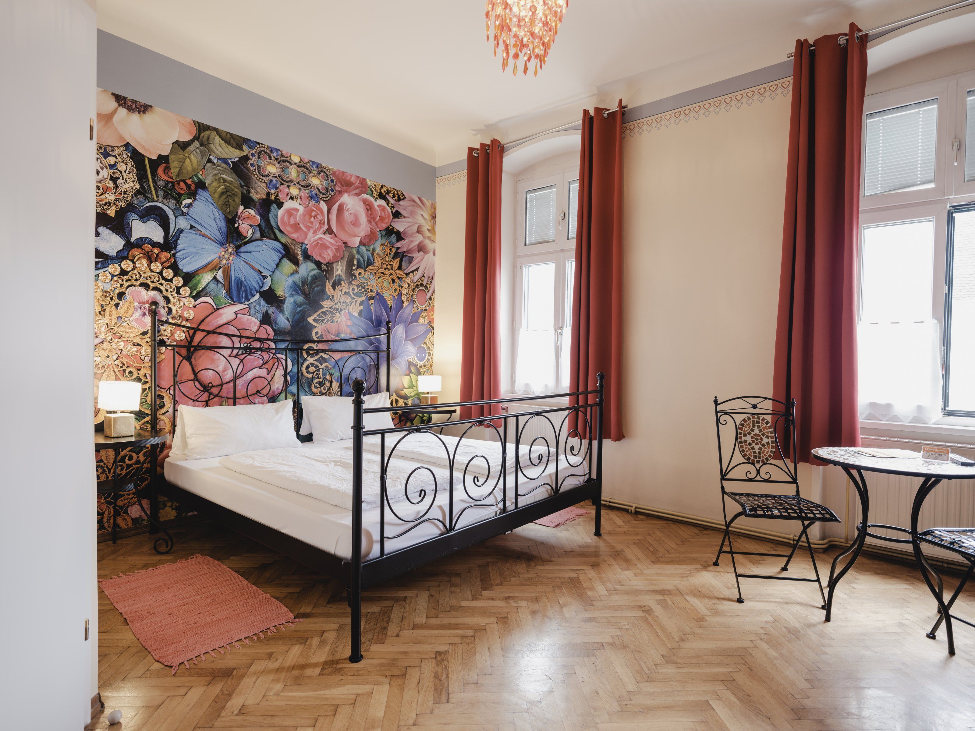 Ein stilvoll eingerichtetes Zimmer mit einem schmiedeeisernen Bett, floraler Wandtapete und einem kleinen Tisch mit Stühlen vor großen Fenstern.