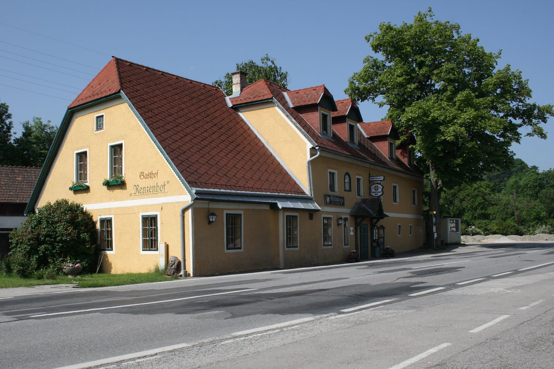 Gelbes Gasthaus mit rotem Dach an einer Straße, umgeben von Bäumen.