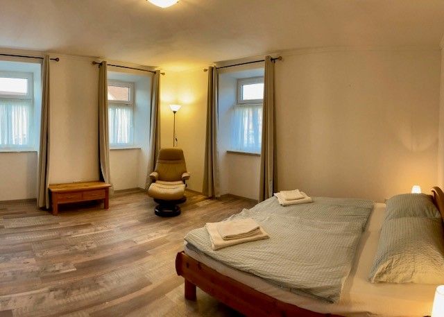 Ein schlichtes Schlafzimmer mit Doppelbett, Sessel, Nachttisch und drei Fenstern mit Vorhängen.