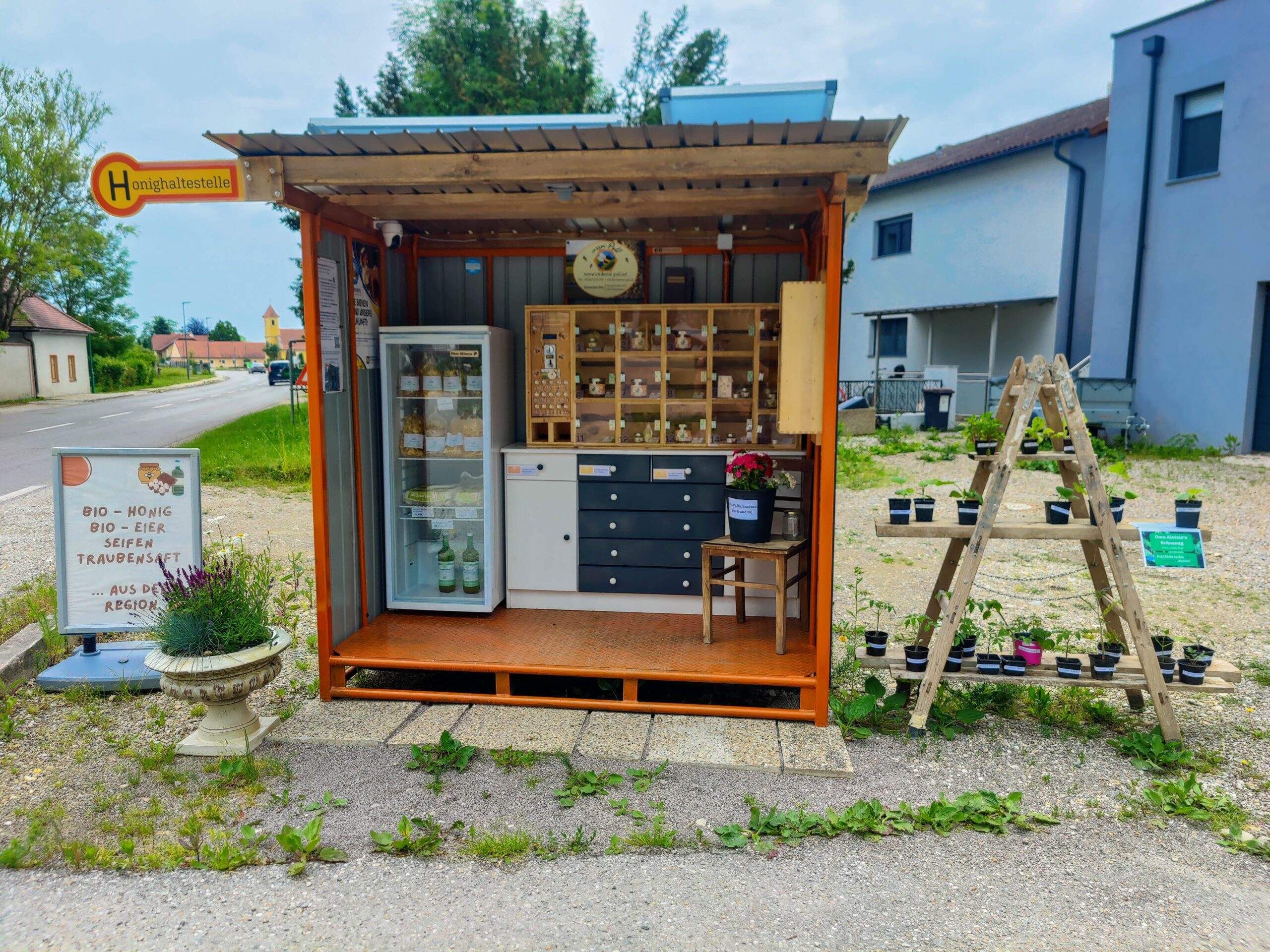 Ein kleiner Verkaufsstand am Straßenrand mit Bio-Produkten wie Honig, Eiern und Seifen. Daneben stehen Pflanzen auf einem Holzgestell.