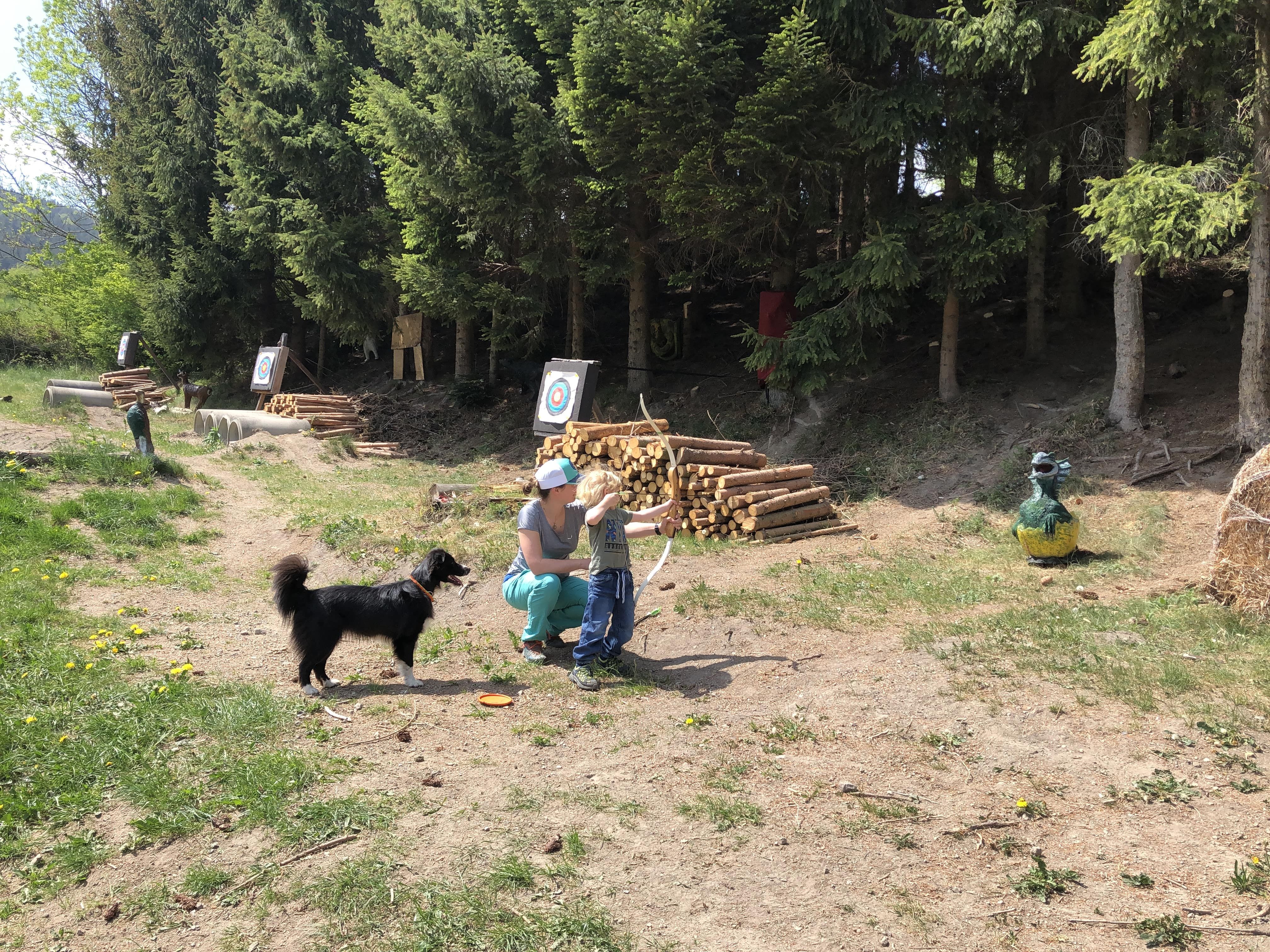 Ein Kind und eine erwachsene Person üben Bogenschießen im Freien, begleitet von einem Hund. Im Hintergrund sind Zielscheiben und Bäume zu sehen.
