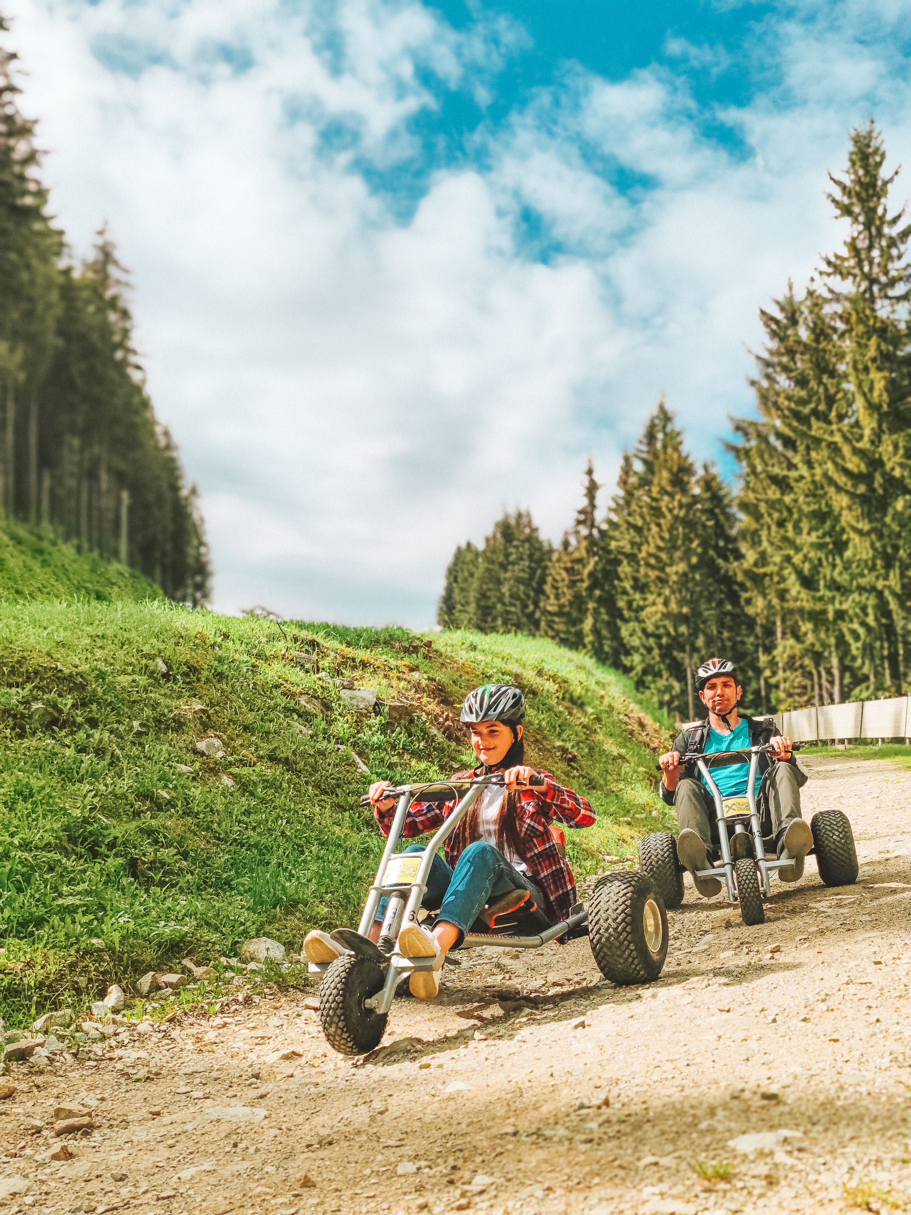Zwei Personen fahren Mountaincarts auf einem Schotterweg, umgeben von Bäumen und blauem Himmel.
