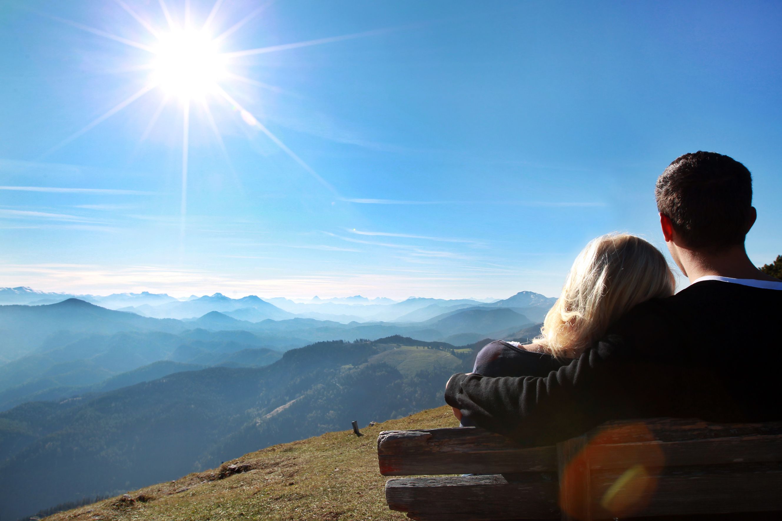 Ein Paar sitzt auf einer Bank und blickt auf eine Berglandschaft unter einem strahlend blauen Himmel mit Sonne.