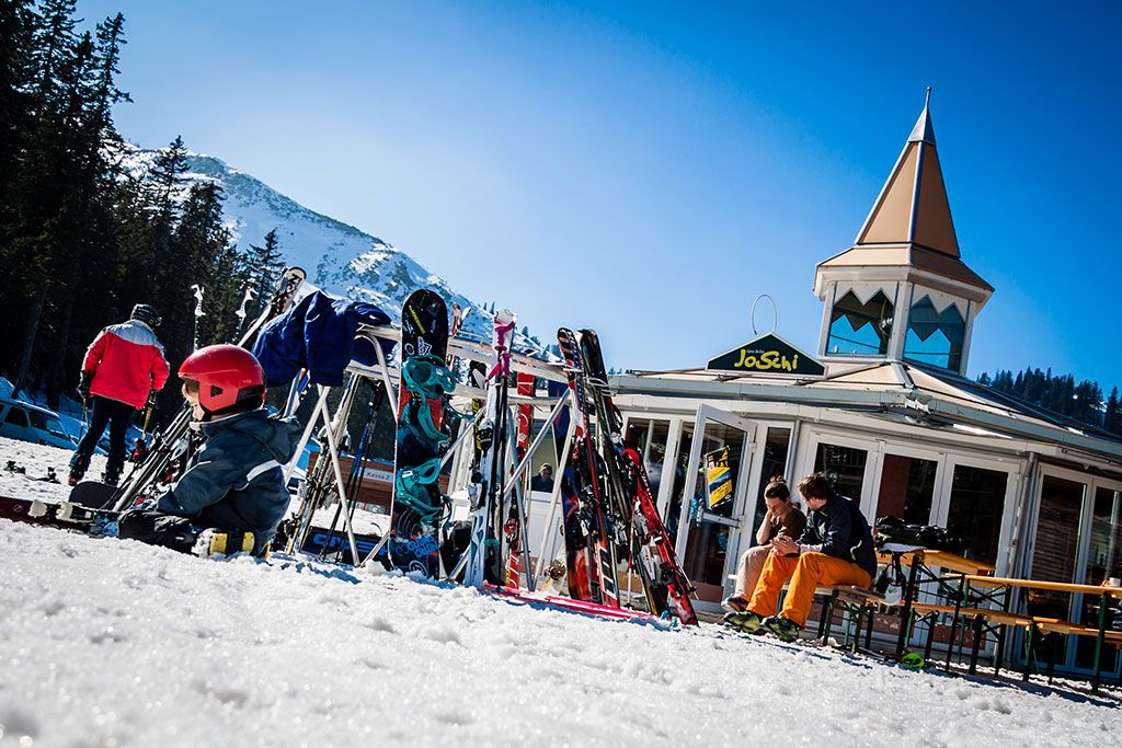 Skifahrer vor der JoSchi-Bar mit Skiern und Snowboards im Schnee.