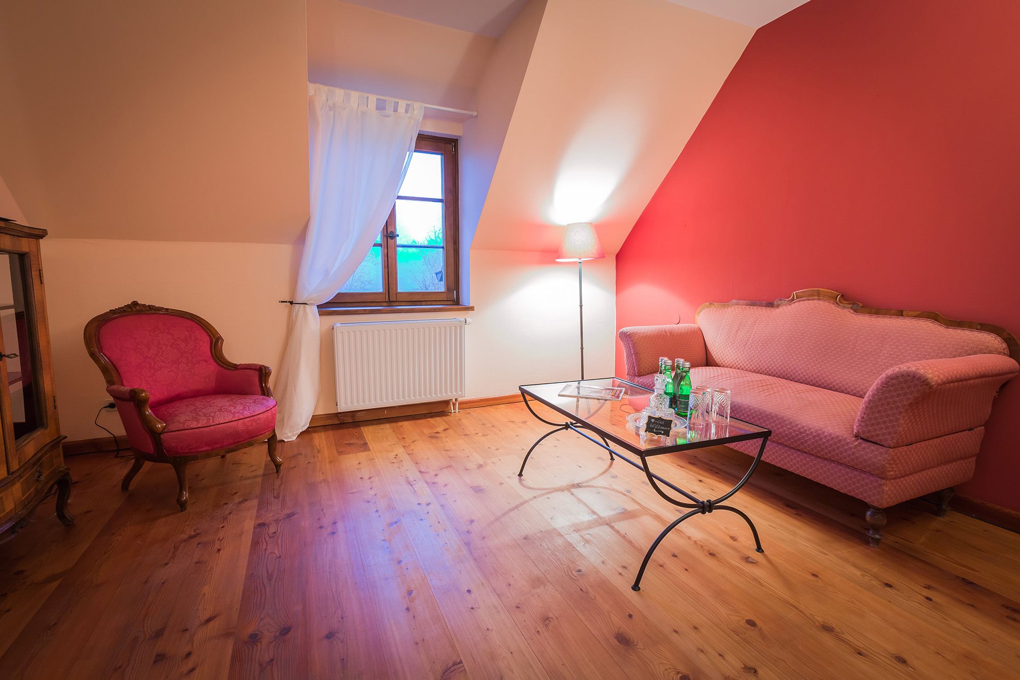 Gemütliches Wohnzimmer mit rotem Sofa, Sessel, Glastisch und Stehlampe vor roter Wand.