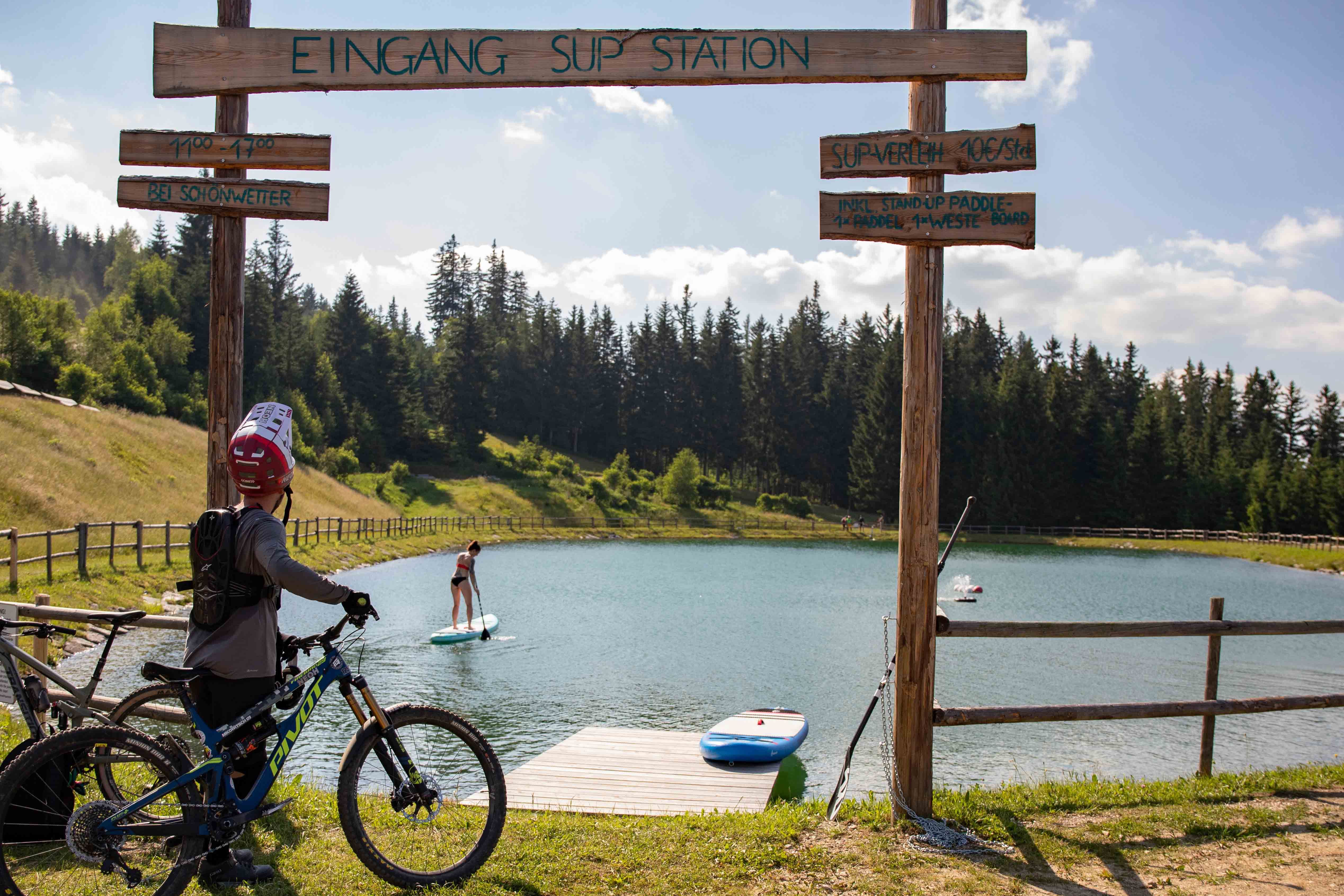 Ein Radfahrer steht vor einem Speicherteich mit SUP-Station in St. Corona am Wechsel. Eine Person paddelt auf dem Teich.