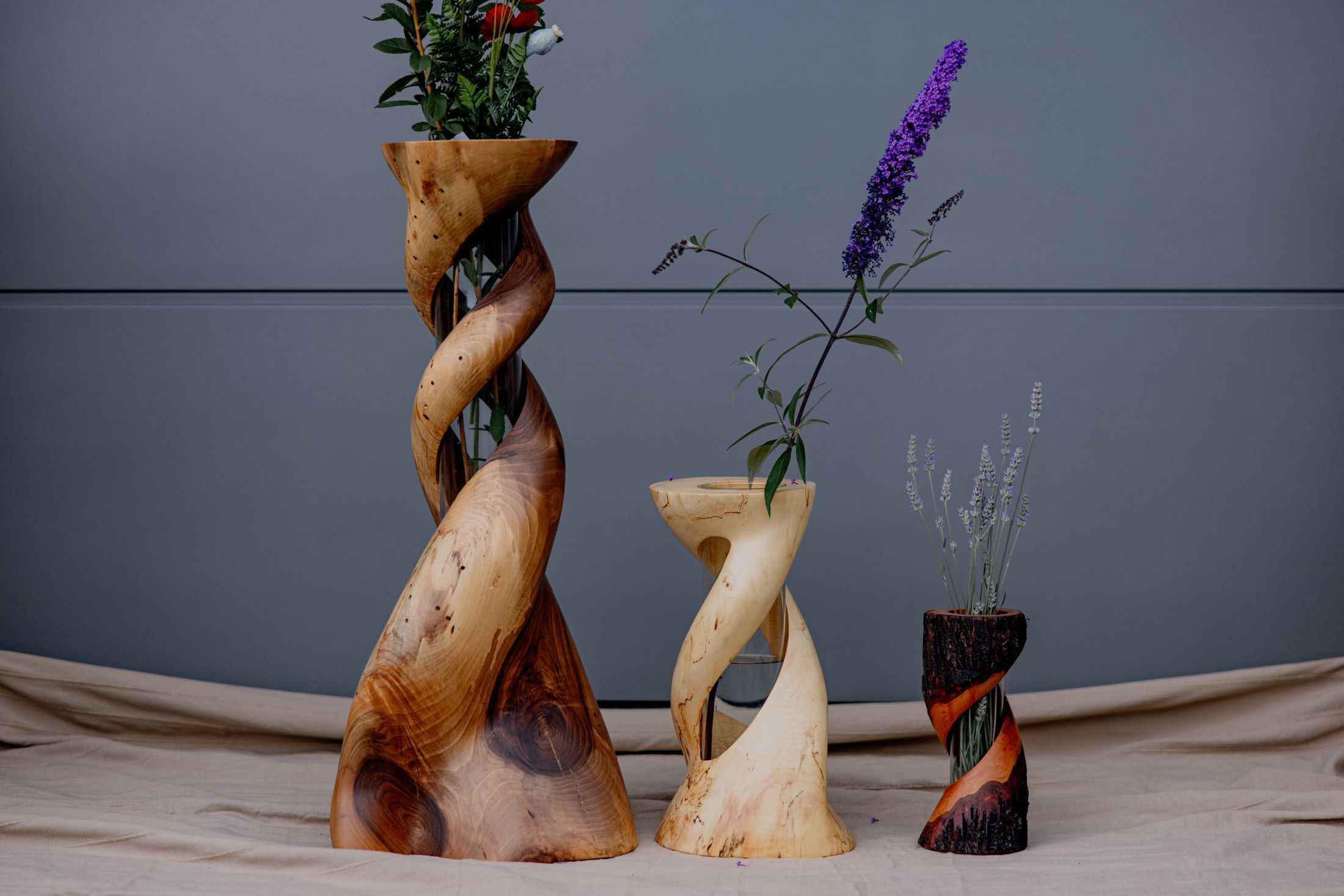 Drei gedrehte Holzvasen mit Blumen vor grauem Hintergrund.