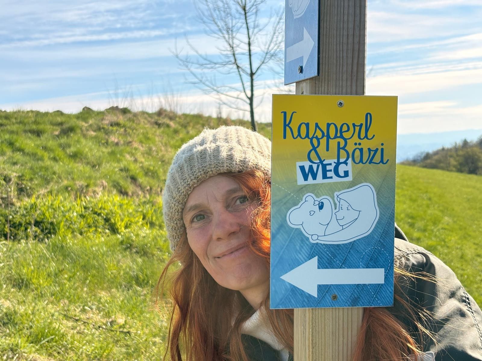 Eine Frau mit Mütze steht neben einem Wegweiser mit der Aufschrift 'Kasperl & Bäzi Weg' in einer grünen Landschaft.