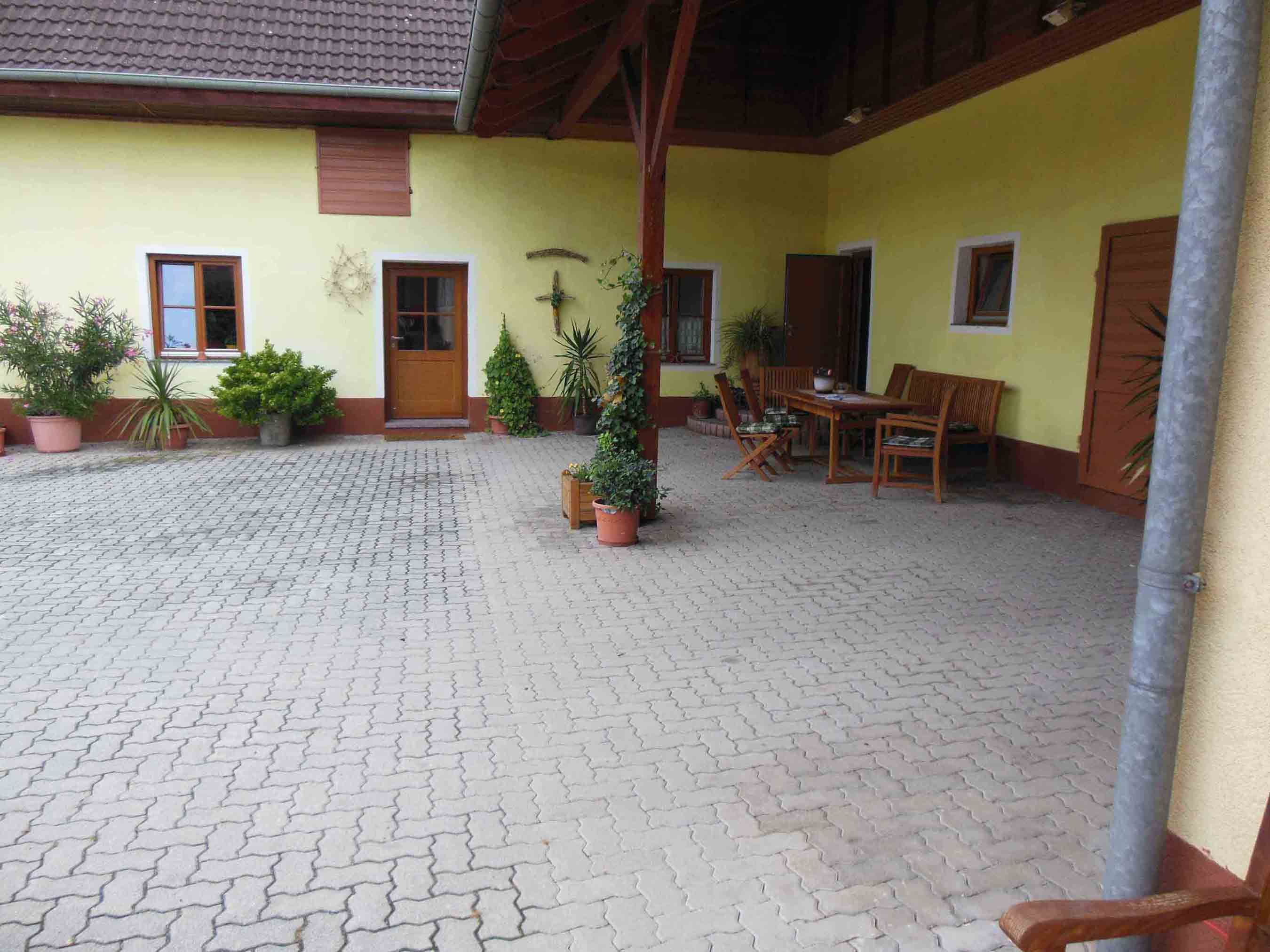 Innenhof mit gelber Fassade, Holztisch und Stühlen, umgeben von Pflanzen in Töpfen.