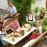 Eine Familie in traditioneller Kleidung sitzt an einem Tisch im Garten und bereitet eine Mahlzeit vor.
