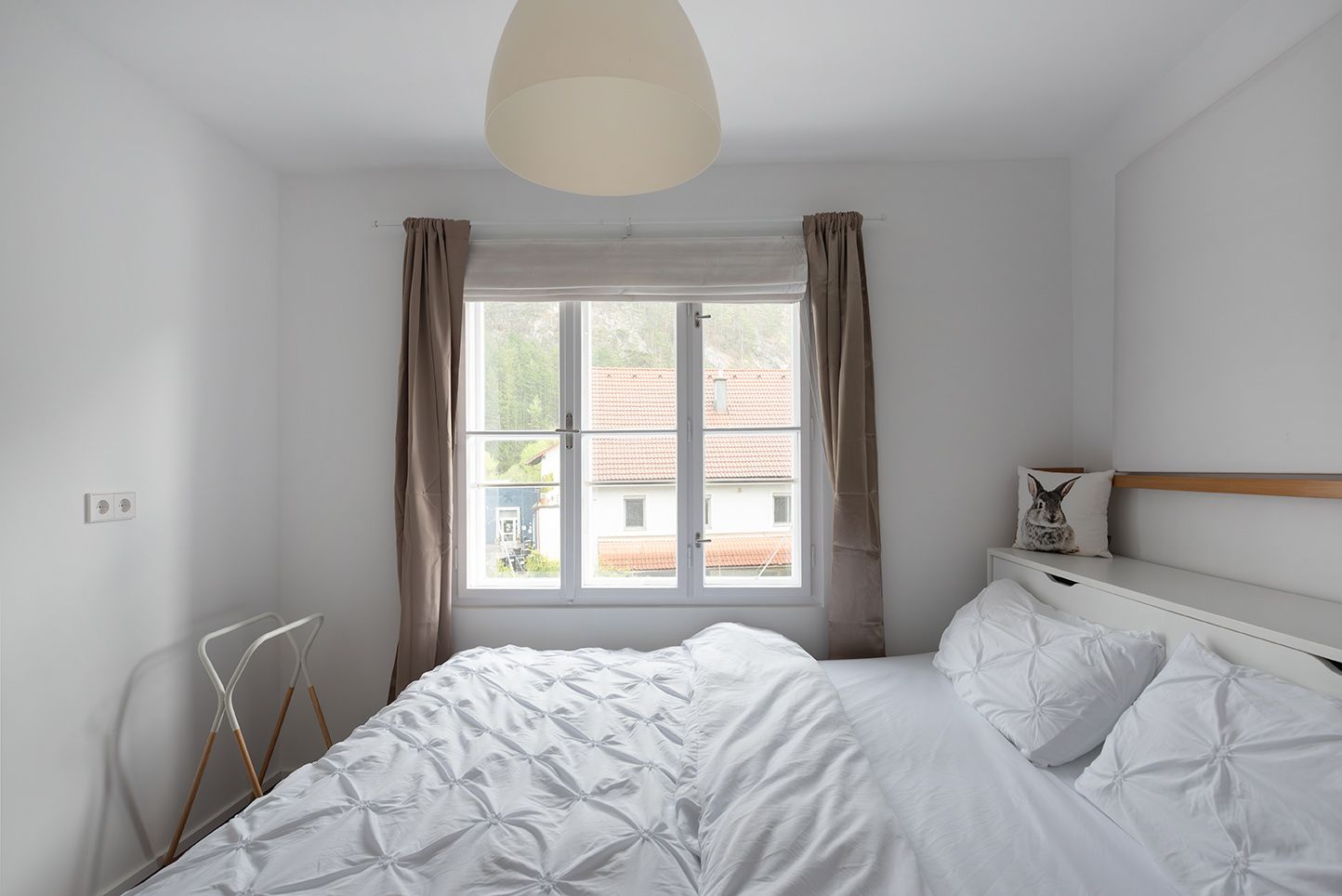 Helles Schlafzimmer mit weißem Bett, Fenster mit Blick auf ein Hausdach und braunen Vorhängen.